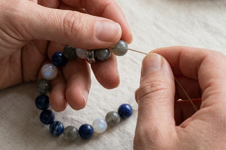 Deux mains montant un bracelet a la main, perle par perle, sur un plan de travail en lin - visuel provisoire