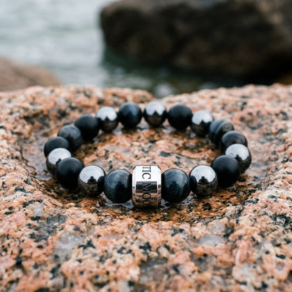 Bracelet Le Rempart en onyx, hematite et oeil de faucon, pose dans un creux de granit rose sur la cote bretonne - visuel provisoire